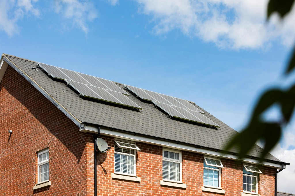 A red brick house with solar panels installed on its roof under a partly cloudy sky.