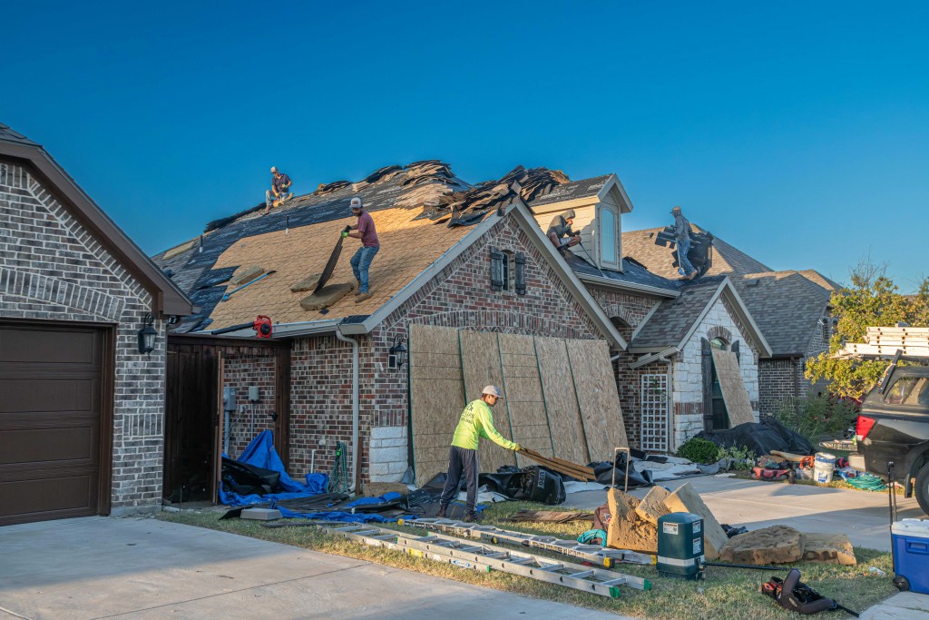 Roofing contractors replacing shingles and installing new roofing materials on a brick residential house under clear blue sky