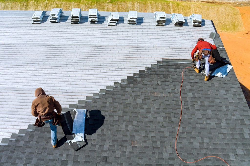 An aerial view of two construction workers in safety gear installing new gray asphalt roof shingles, contrasting with the light underlayment on the adjacent section of the roof.