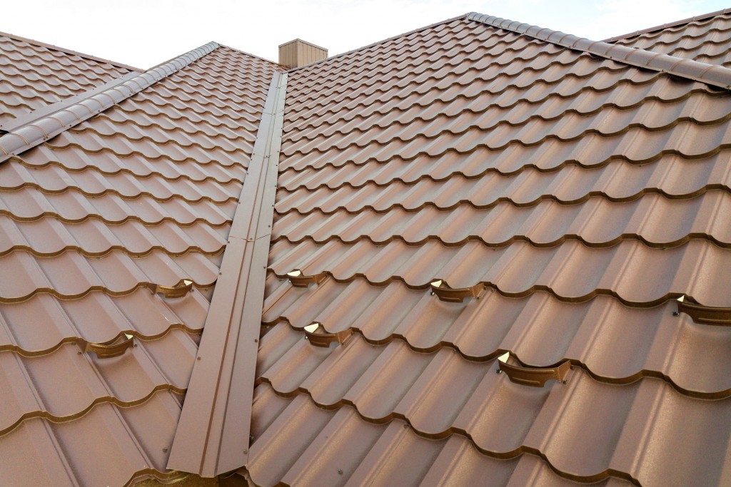 Brown metal roofing with tile-profile panels, showing ridge caps and valley flashing on a residential roof.