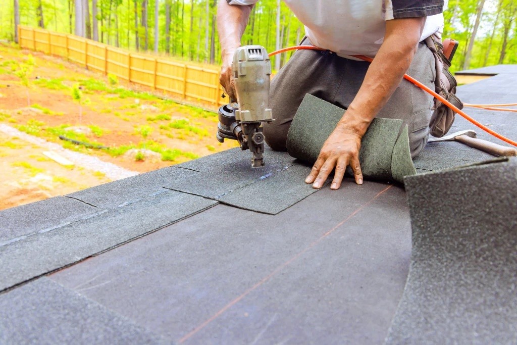 Worker installing asphalt shingles on a roof using a pneumatic nail gun, with roofing materials laid out during construction.