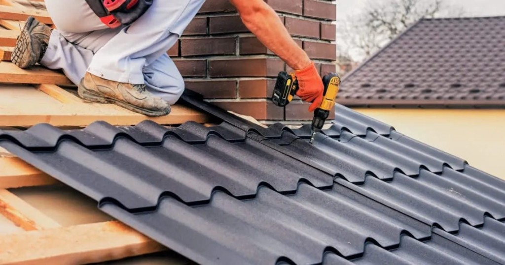 A roofer wearing orange gloves uses a power drill to fasten dark gray metal roofing panels near a brick chimney, with wooden roof decking and a neighboring shingled roof visible in the background.